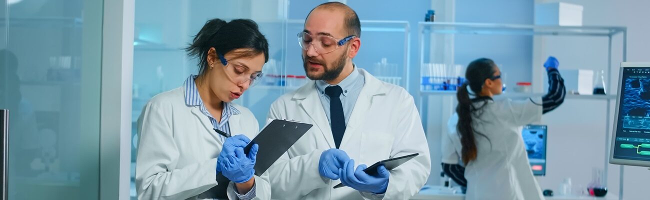 Group of medical researchers discussing about vaccine development standing in equipped lab pointing on tablet and taking notes. Doctors examining virus evolution using high tech researching diagnosis