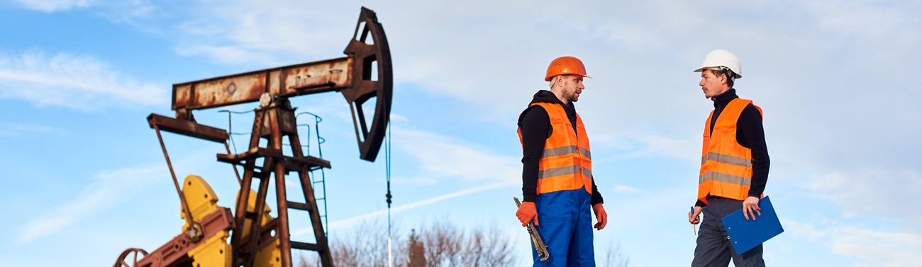 Two oil men in helmets and work vests standing near oil well pump jack and discussing work. Oil worker holding clipboard and talking with colleague at oil field. Concept of petroleum industry.